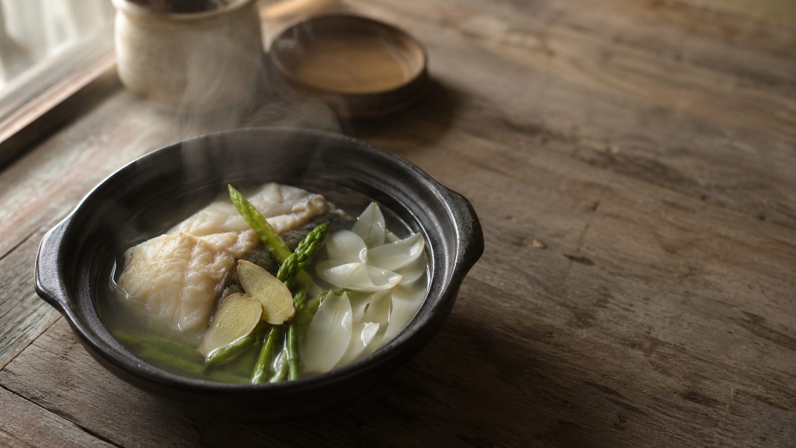 A clay pot of clear broth with white fish, asparagus, lily bulb, and ginger, photographed in soft window light.